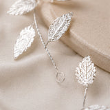A close-up of a silver leaf vine or garland with detailed leaf motifs and twisted stems, arranged on a soft beige fabric surface.