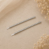 Two slim silver hair claw clips embellished with clear rhinestones, arranged diagonally on a textured beige surface with soft cream fabric and dried flowers in the corners.