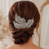 A silver feather-shaped hair accessory embellished with small clear rhinestones, pinned to the back of a woman's styled brown hair.