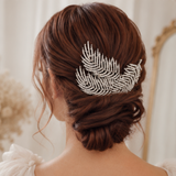 A woman shown from the back wearing a brown updo hairstyle with a silver feather-shaped hair accessory adorned with small clear rhinestones, against a soft cream indoor background.
