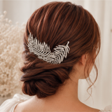 A brown hairpiece accessory styled in a woman's low bun, viewed from the back. The hair accessory is a silver-toned leaf-shaped hair clip embellished with small clear rhinestones, set against a soft cream background.