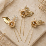Four gold-toned hair pins styled with floral and leaf motifs, pearls, and rhinestones, arranged on a textured beige stone surface with dried flowers.