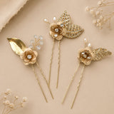Four gold-toned floral hair pins with pearl and crystal accents, arranged on a warm beige background with dried floral props.