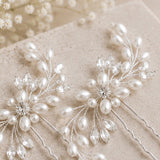 Two bridal hair pins decorated with faux pearls, clear crystal beads, and silver-toned wire branches, arranged on a textured beige surface with soft baby's breath flowers in the background.