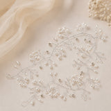 A delicate bridal hair vine featuring silver-tone wire branches adorned with small pearls, clear rhinestones, and bead accents, laid flat on a warm beige fabric background with soft cream tulle and dried flowers.