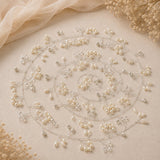 A circular arrangement of pearl-like beads, clear rhinestones, and delicate silver-toned wire on a warm beige fabric background with soft draped fabric and dried floral props.