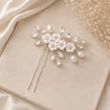 A bridal hairpin featuring white faux-pearl flowers, clear rhinestone leaves, and pearl beads on a silver-tone wire, styled on a beige textured surface with satin and dried floral props.