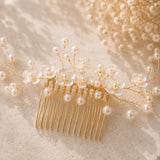 A gold hair comb decorated with white faux pearls and clear glass flowers, styled on a textured beige surface with dried baby's breath.