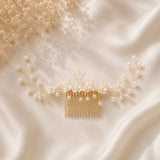 A gold hair comb decorated with delicate pearl-like beads and small floral wire details, arranged on a soft cream satin fabric with dried baby's breath in the corner.