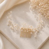 A delicate gold hair comb featuring small pearl-like beads and branching gold wire details, styled on a textured cream stone slab with soft satin fabric and dried baby's breath in the background.