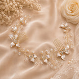 A bridal hair comb accessory with delicate gold-tone wire branches, clear pearl-like beads, and white floral embellishments, arranged on champagne satin fabric with dried flowers and a rose.