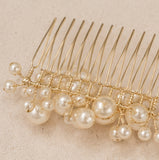 A gold metal hair comb decorated with multiple strands of faux pearl beads in cream and white, resting on a textured beige surface.