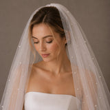 A woman wearing a bridal veil with small pearl-like beads and a strapless white wedding dress, photographed against a plain taupe studio background.