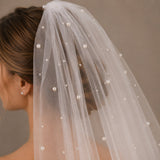 A close-up of a bridal veil with delicate pearl beads woven through sheer white tulle, worn by a model with her hair styled in an updo.