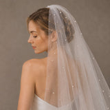 A woman in a white wedding dress shown from the back, wearing a long sheer tulle bridal veil adorned with small pearl-like beads, against a plain taupe studio background.