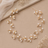 A gold vine necklace with gold leaves and white pearls, styled on a beige background with a draped fabric and dried flowers.