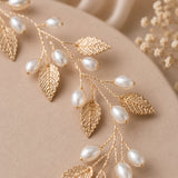A gold-toned leaf vine accessory with dangling white faux pearls, arranged on a soft beige fabric background with dried floral decor in the corner.