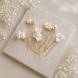 Five white floral hair pins with pearl accents and gold-tone wire stems, arranged on a square beige stone tile with small white flowers around it.
