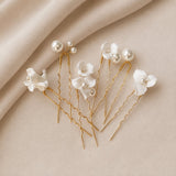 Five white floral hair pins with pearl-like beads and gold-tone wire backs, arranged on a soft beige fabric background.