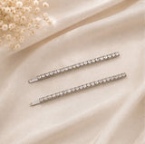 Two slim silver hair claw clips embellished with clear rhinestones, arranged diagonally on a draped cream satin fabric with dried baby's breath flowers in the top left corner.