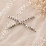 A silver-toned hair barrette with a row of clear rhinestones, arranged in an X shape on a beige lace fabric background with dried baby's breath flowers.