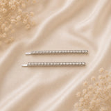 Two silver barrette hair clips embellished with clear crystals, arranged horizontally on a beige satin fabric background with dried baby's breath flowers in the corners.