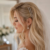 A blonde bride wearing a delicate double-row pearl hair clip and matching pearl drop earrings, shown in profile with a white lace wedding dress and veil.