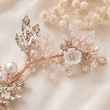 A delicate floral hair claw clip in rose gold, featuring a white flower, pink beads, clear crystals, and faux pearls, styled on a soft beige fabric background.