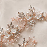 A close-up of a delicate bridal hairpiece featuring rose gold wire branches adorned with white pearl flowers, white pearls, clear rhinestones, and pale pink bead clusters, laid on a soft beige fabric background.
