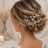 A woman shown from the back wearing a delicate bridal hairpiece with white pearls and rose gold floral branches tucked into her styled updo. She is wearing a white lace wedding dress and matching pearl drop earrings in a softly lit, elegant setting.