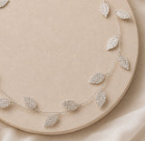 A delicate silver leaf garland draped over a round beige display board on a soft neutral fabric background.