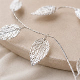 A silver leaf garland with textured, detailed leaf pendants draped over a beige fabric surface.
