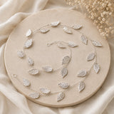 A delicate silver leaf garland draped in a spiral pattern on a round beige stone tray, set against a background of soft beige fabric and dried flowers.