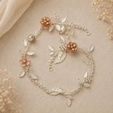 A delicate floral vine headpiece in silver-toned metal with pale pink flowers and clear rhinestone accents, arranged on a soft beige fabric background with dried floral props.