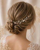 A close-up back view of a woman with styled brown hair wearing a delicate floral and leaf hairpiece in soft pink, champagne, and white tones, set against a warm cream background.