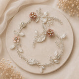 A delicate floral and leaf jewelry piece arranged in a circular flat lay on a round beige stone plate, with soft beige fabric and dried flowers around it. The item appears to be a silver-toned vine-style accessory or headpiece with pale blush pink flowers and clear rhinestone-like accents.