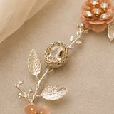 A close-up of a decorative floral hair accessory with silver-toned leafy branches, peach fabric flowers, and a faceted clear central stone, styled on a beige textured surface with soft fabric in the corner.