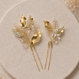Two gold leaf and pearl hair pins displayed on a round beige stone plate with dried floral accents in the corners.