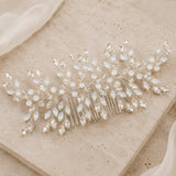 A silver bridal hair comb decorated with clear faceted crystals and milky white iridescent beads, styled on a textured beige stone tile.