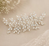 A silver bridal hair comb decorated with clear crystals and iridescent white rhinestones, styled on a beige fabric background with baby's breath flowers and a pearl-trimmed veil.